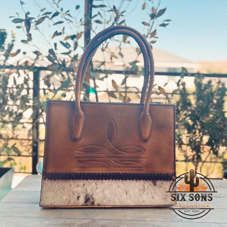 Puede incluir: Un bolso de cuero marr&oacute;n con dos asas, un patr&oacute;n decorativo cosido en la secci&oacute;n superior y un panel de piel de vaca texturizada en la base con un borde trenzado oscuro. El logotipo Six Sons Mercantile Est. 2021 es visible en la esquina.