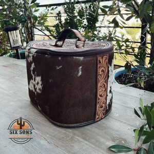 May include: A brown and white cowhide leather travel case with a tooled leather trim and a handle. The case is closed and has a zipper.