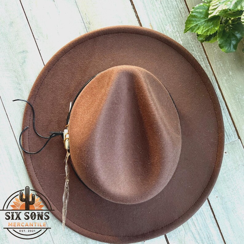 May include: A brown felt fedora hat with a brown leather band and a feather accent. The hat has a wide brim and a tall crown.