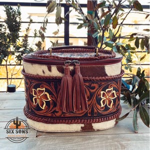 May include: A round, brown and white leather cosmetic bag with a floral design and tassels. The bag has a zipper closure and a top handle. The bag is made by Six Sons Mercantile.