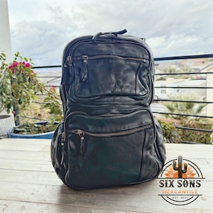 May include: Black leather backpack with multiple zippered compartments. The bag has a rounded top and a rectangular bottom. The backpack is sitting on a wooden surface. The logo for Six Sons Mercantile is visible in the bottom right corner.