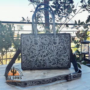 May include: A black leather handbag with a floral tooled design and a detachable shoulder strap. The bag has a top handle and a flap closure. The strap is also tooled with a floral design.