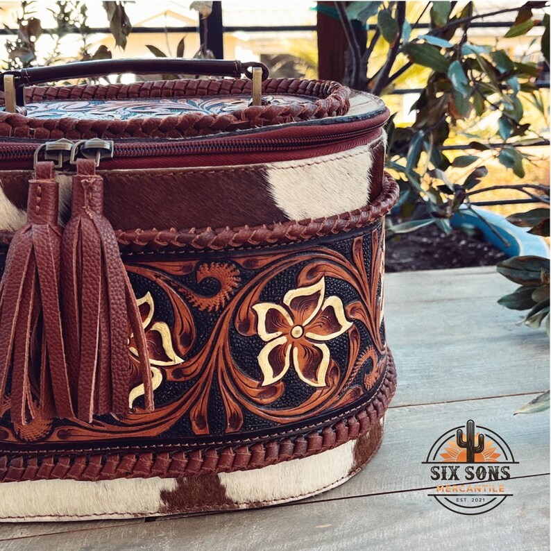 May include: A brown leather oval-shaped handbag with a braided handle and a tassel. The bag is decorated with intricate floral tooling and a white and brown cowhide pattern. The bag is labeled "Six Sons Mercantile Est. 2021".