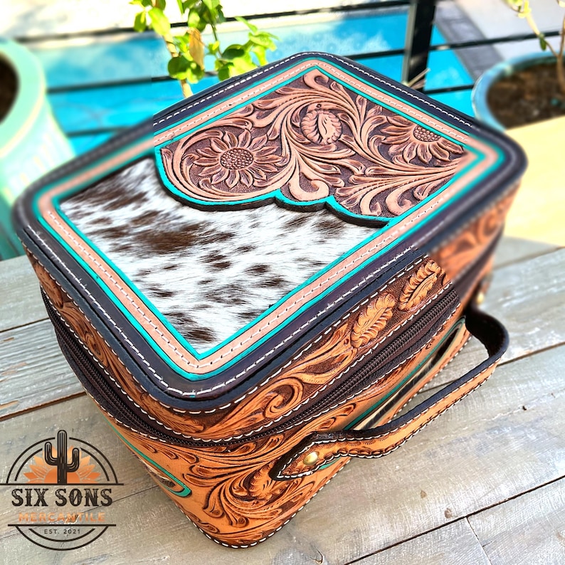 May include: A handcrafted leather travel case with a cowhide top and floral tooling. The case features a turquoise border, a brown zipper, and a handle. The brand logo "SIX SONS" is visible on the bottom left.
