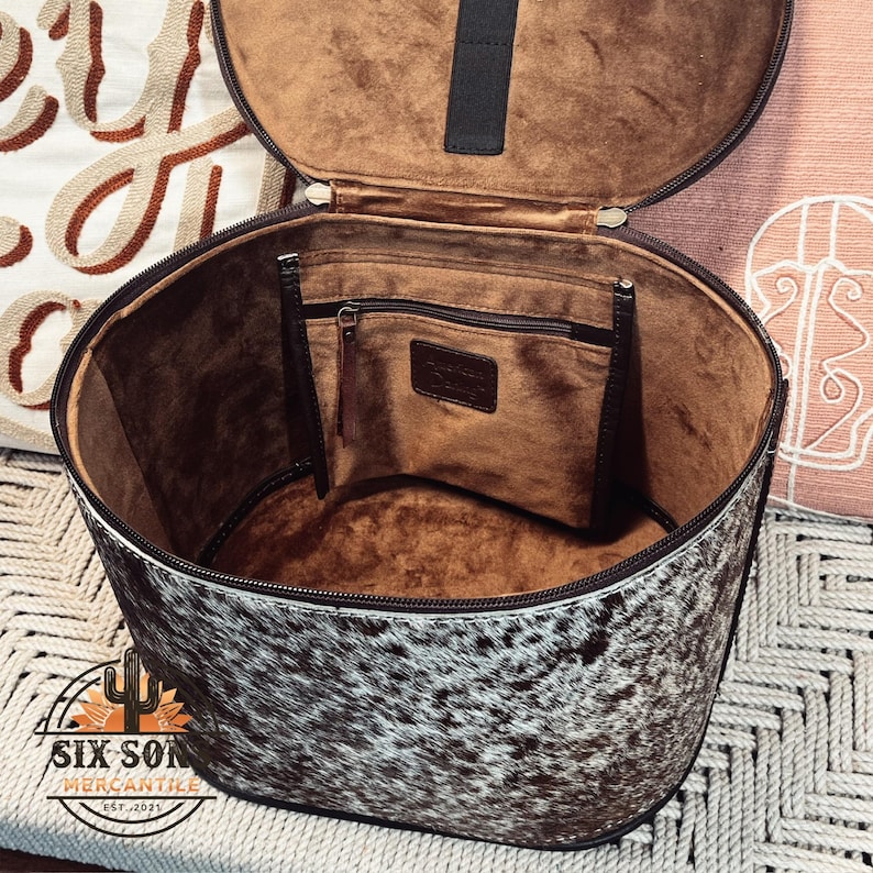 May include: A white and black cowhide round travel bag with a brown leather interior. The bag has a zippered pocket on the inside and a brown leather handle.