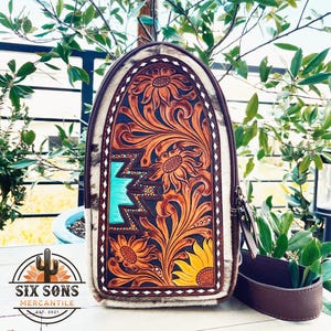 May include: A brown leather crossbody bag with a white and brown cowhide pattern. The bag features a turquoise and brown floral design with sunflowers. The bag has a brown leather strap.
