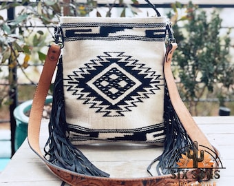 Puede incluir: Un bolso bandolera blanco y negro con estampado geométrico, con una correa de cuero marrón y flecos negros. La correa tiene un diseño floral. El bolso tiene el texto "Six Suns Mercantile Est. 2021" en la parte inferior.