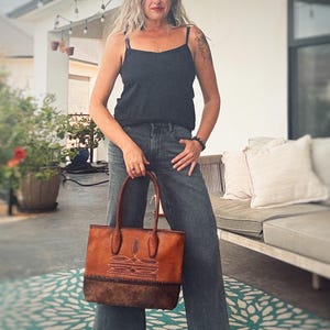 Puede incluir: Un bolso tote rectangular de cuero marr&oacute;n con un patr&oacute;n de costuras decorativas en la parte superior y una base de cuero marr&oacute;n oscuro texturizado con un borde trenzado, equipado con dos asas redondeadas y resistentes.
