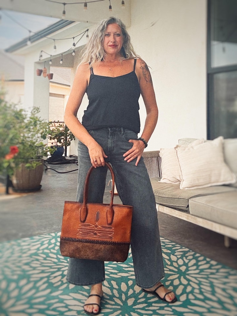 Puede incluir: Un bolso tote rectangular de cuero marr&oacute;n con un patr&oacute;n de costuras decorativas en la parte superior y una base de cuero marr&oacute;n oscuro texturizado con un borde trenzado, equipado con dos asas redondeadas y resistentes.