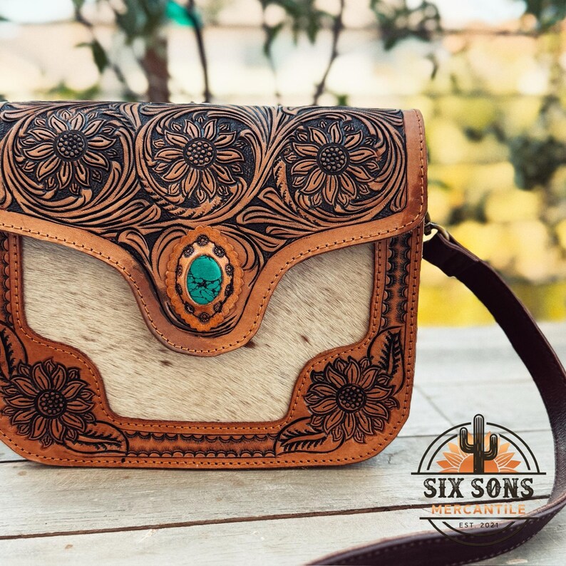 May include: A brown leather crossbody bag with a floral sunflower design and a turquoise stone accent. The bag features a light-colored hair-on hide panel and a dark brown strap. The logo "SIX SONS MERCANTILE EST. 2021" is visible.