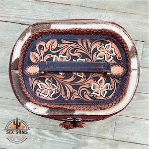 May include: A brown leather oval-shaped vanity case with a tooled floral design. The case has a brown leather handle and a white and brown cowhide exterior.