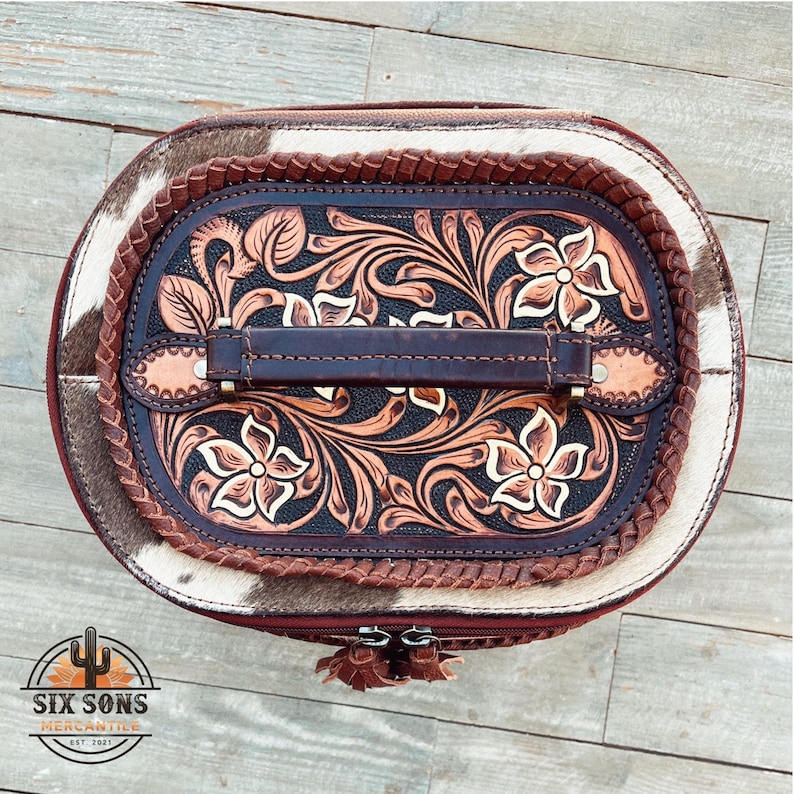 May include: A brown leather oval-shaped vanity case with a tooled floral design. The case has a brown leather handle and a white and brown cowhide exterior.