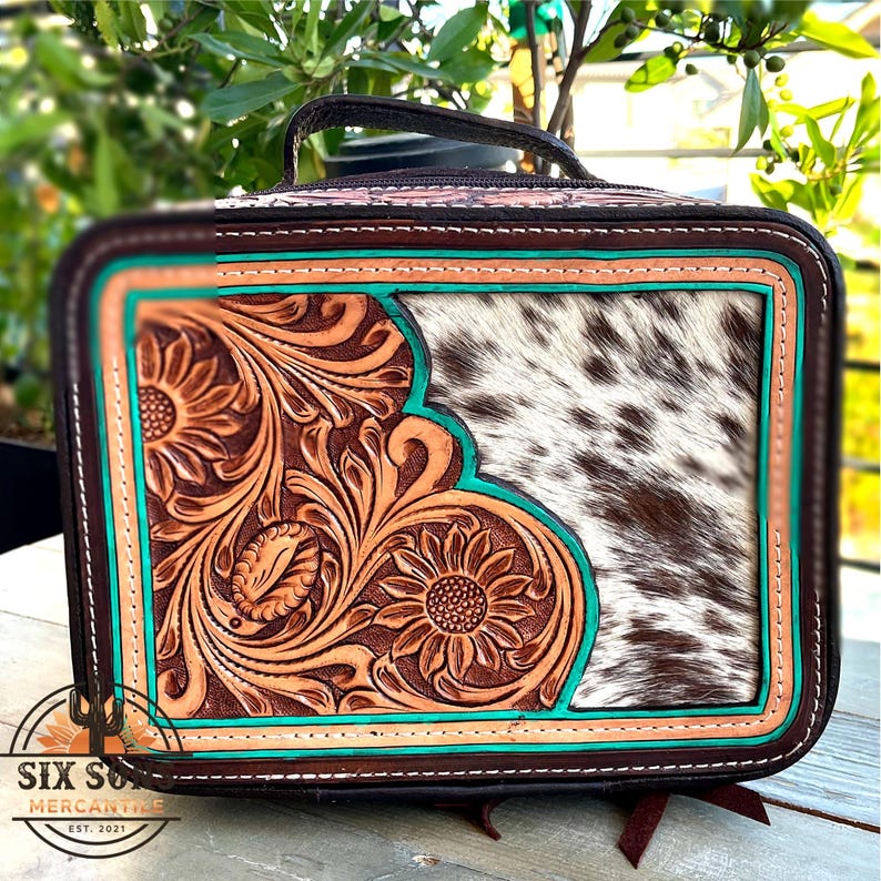 May include: A rectangular, brown leather bag with a handle, featuring a floral tooled design in tan and turquoise accents. The right side has a cowhide panel. The bag has the text "SIX SUNS MERCANTILE EST. 2021".