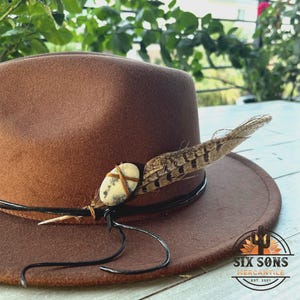 May include: A brown felt fedora hat with a black leather band and a feather accent. A small, white stone with brown accents is attached to the band. The hat has a logo on the side that reads "Six Sons Mercantile Est. 2021".