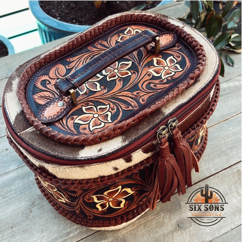 May include: A brown leather oval-shaped vanity case with a floral tooled design. The case has a brown leather handle and a zipper closure with a brown tassel. The case is made by Six Sons Mercantile, est. 2021.
