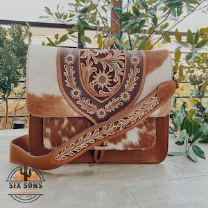 May include: A brown and white cowhide leather crossbody bag with a floral embossed flap and strap. The bag features a brown leather strap and accents, with a buckle closure. The bag has a front pocket and the logo "SIX SONS MERCANTILE EST. 2021".