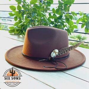 May include: A brown felt fedora hat with a black band and a feather accent. A small white stone is tied to the feather with twine. The hat is sitting on a white wooden surface.