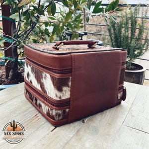 May include: A brown leather and cowhide two-tiered box with a gold handle. The box has a zipper closure and is perfect for storing jewelry or other small items.