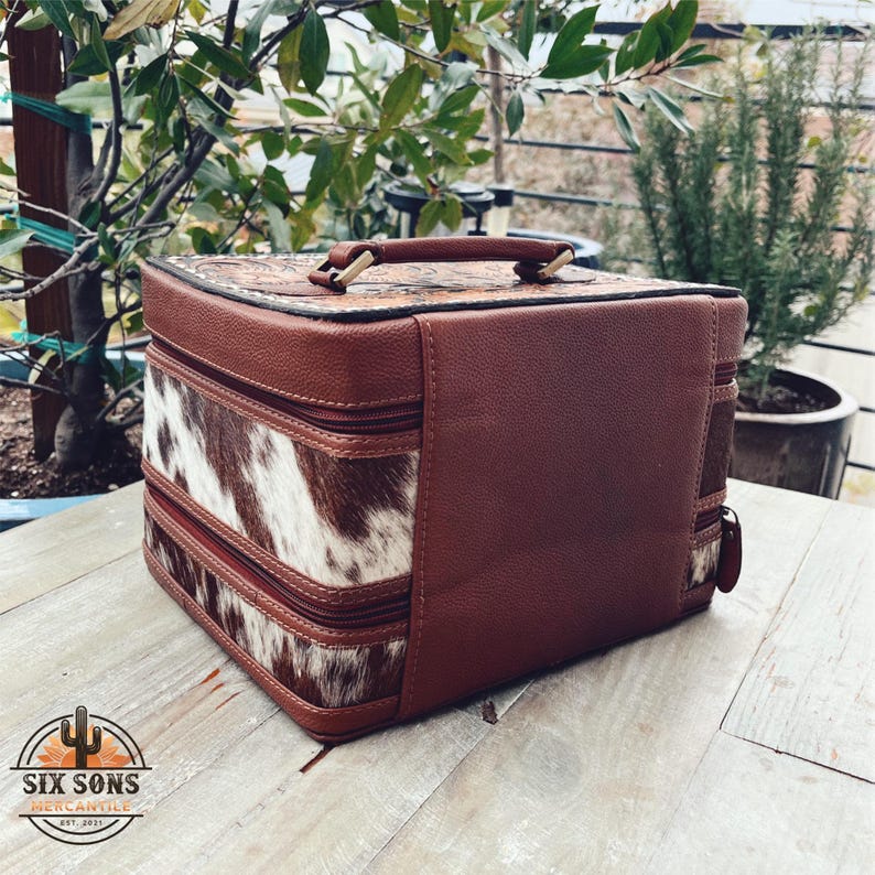 May include: A brown leather and cowhide two-tiered box with a gold handle. The box has a zipper closure and is perfect for storing jewelry or other small items.