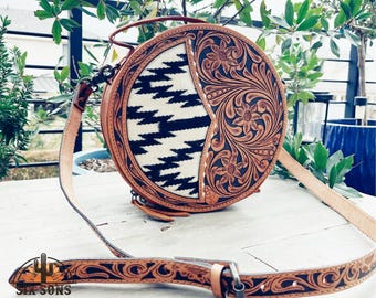 May include: A round, brown leather purse with a black and white geometric pattern on one side and a floral design on the other side. The purse has a long, adjustable strap.