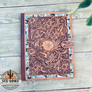 May include: A brown leather journal with a tooled floral design and a turquoise and white geometric border. The journal has a brown leather strap with a metal buckle.