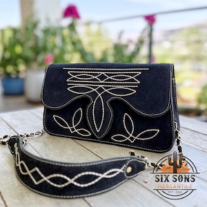May include: Black suede crossbody bag with white stitching and a decorative design on the flap. The bag features a chain strap and a matching suede shoulder strap. The bag has the text "SIX SONS MERCANTILE EST. 2021".
