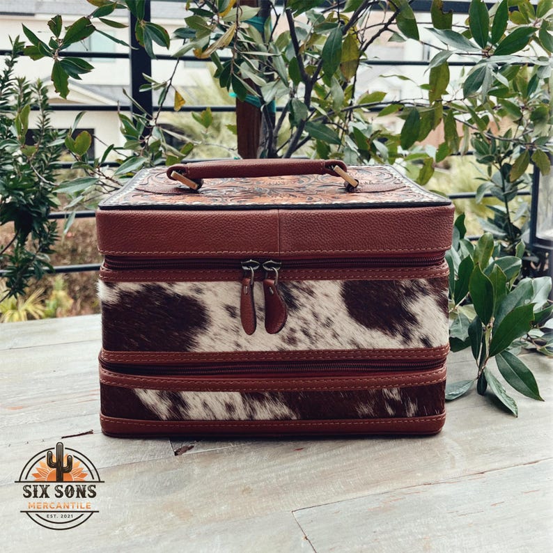 May include: A brown leather and cowhide two-tiered vanity case with a floral tooled design on the top. The case has a handle and two zippers.