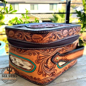 May include: A brown leather bag with intricate floral tooling and a zipper closure. The bag features a turquoise accent and a handle. The text "SIX SONS MERCANTILE EST. 2021" is visible on the bottom left.
