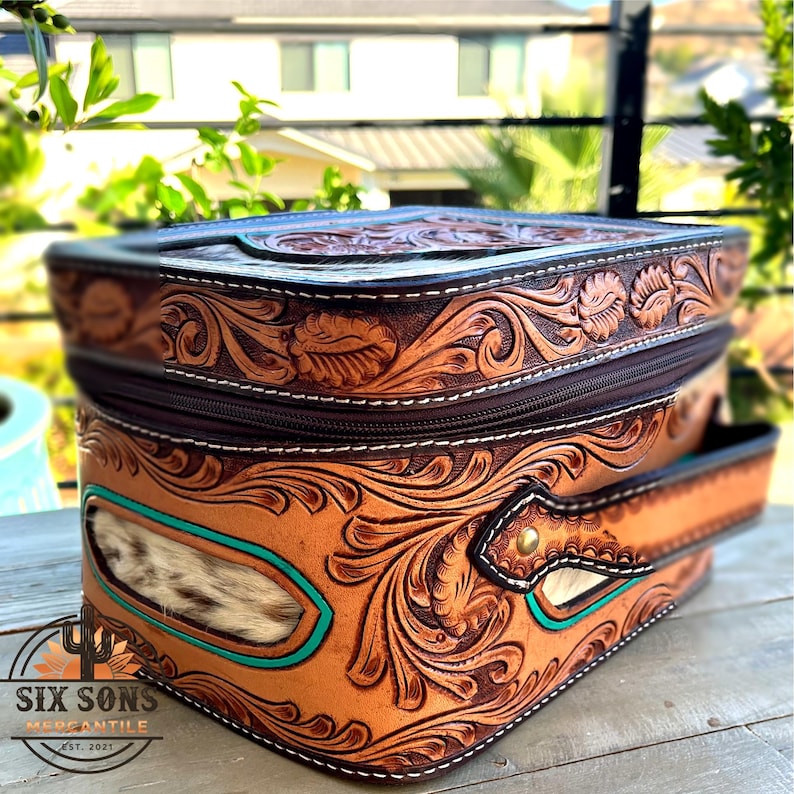 May include: A brown leather bag with intricate floral tooling and a zipper closure. The bag features a turquoise accent and a handle. The text "SIX SONS MERCANTILE EST. 2021" is visible on the bottom left.