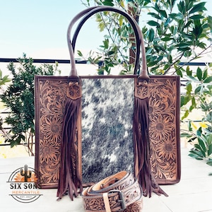 May include: A brown leather tote bag with a cowhide panel and floral tooled leather accents. The bag has fringe detailing and a matching belt with a buckle.