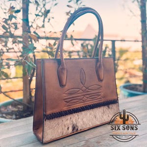 Puede incluir: Un bolso tote de cuero marr&oacute;n con dos asas, un patr&oacute;n decorativo cosido en la secci&oacute;n superior y un panel de piel de vaca texturizada en la parte inferior con un borde trenzado oscuro. El logotipo SIX SONS MERCANTILE EST. 2021 es visible.
