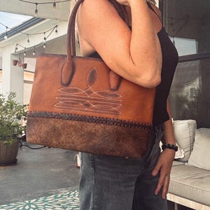 Puede incluir: Un bolso tote de cuero color canela con una base de cuero texturizado marr&oacute;n oscuro y un detalle de borde trenzado. La parte superior del bolso muestra costuras blancas decorativas en un patr&oacute;n sim&eacute;trico e incluye dos asas de cuero resistentes.