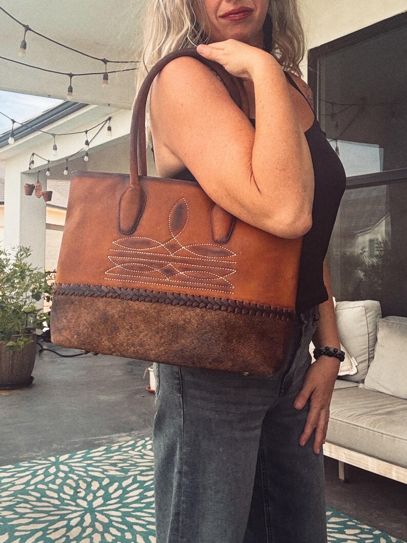 Puede incluir: Un bolso tote de cuero color canela con una base de cuero texturizado marr&oacute;n oscuro y un detalle de borde trenzado. La parte superior del bolso muestra costuras blancas decorativas en un patr&oacute;n sim&eacute;trico e incluye dos asas de cuero resistentes.