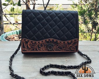 Puede incluir: Bolso bandolera de cuero negro acolchado con un panel inferior de cuero repujado con motivos florales. El bolso tiene una correa de cadena y un cierre de metal. El bolso se muestra sobre una superficie de madera.