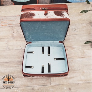 May include: Open brown and white cowhide leather travel case with a blue interior and black elastic straps. The case has a zipper closure.