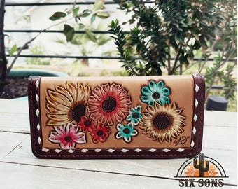 Puede incluir: Una cartera de cuero marrón con un diseño floral que presenta girasoles, margaritas y otras flores en tonos de rosa, amarillo, azul y verde. La cartera tiene un borde cosido marrón y un detalle de costura blanca.