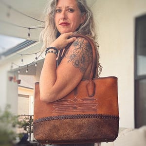 Puede incluir: Un bolso tote de cuero marr&oacute;n con una secci&oacute;n superior lisa con costuras blancas decorativas y un panel inferior texturizado de color marr&oacute;n oscuro con un detalle de borde de cuero trenzado. Incluye dos asas de cuero y pies de metal en la base.