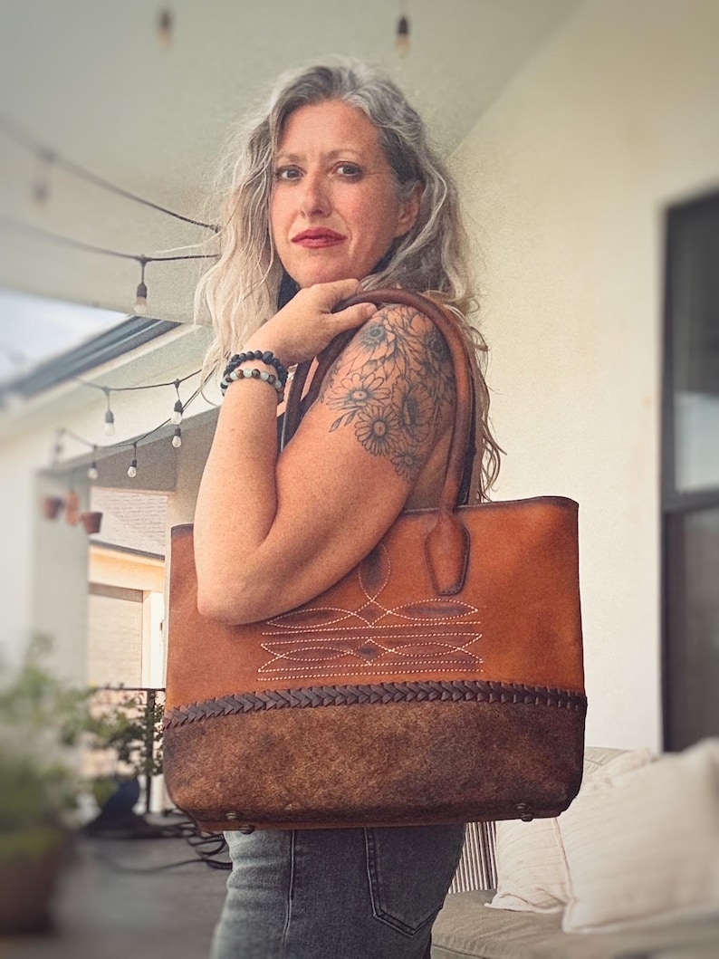 Puede incluir: Un bolso tote de cuero marr&oacute;n con una secci&oacute;n superior lisa con costuras blancas decorativas y un panel inferior texturizado de color marr&oacute;n oscuro con un detalle de borde de cuero trenzado. Incluye dos asas de cuero y pies de metal en la base.