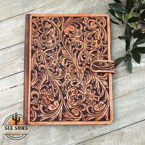 May include: A brown leather journal with a floral tooled design. The cover features a detailed pattern of flowers and vines, with a strap closure. The journal is made of high-quality leather and has a rustic, handcrafted look.
