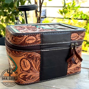 May include: A brown leather cosmetic bag with a floral tooled design and a black leather panel. The bag has a zipper closure, white stitching, and a turquoise accent. The logo "SIX SON MERCANTILE EST. 2021" is visible.