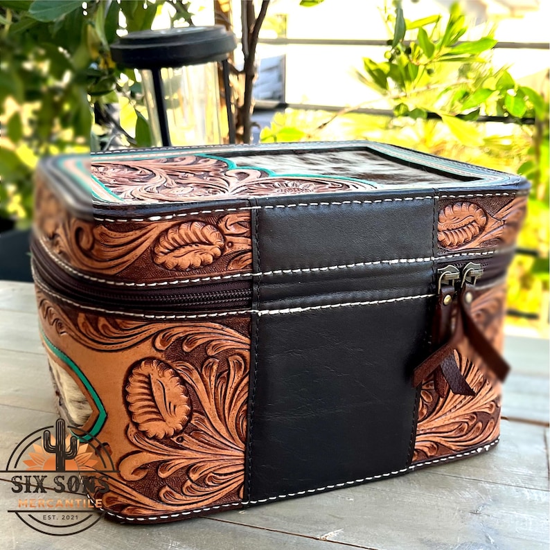 May include: A brown leather cosmetic bag with a floral tooled design and a black leather panel. The bag has a zipper closure, white stitching, and a turquoise accent. The logo "SIX SON MERCANTILE EST. 2021" is visible.