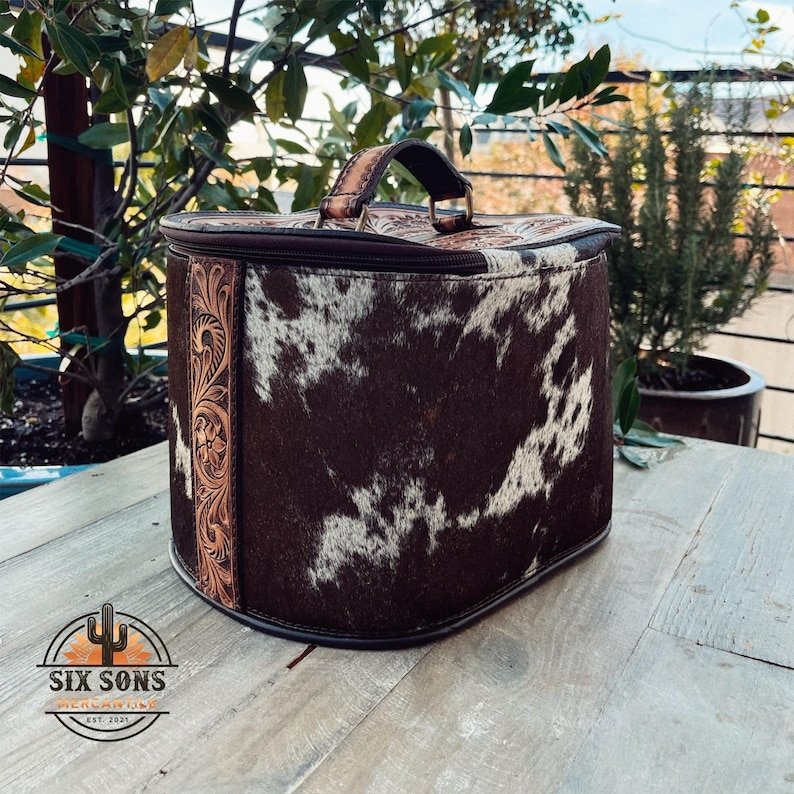 May include: A brown and white cowhide travel case with a tooled leather handle and trim. The case has a zipper closure and is perfect for storing and transporting small items.