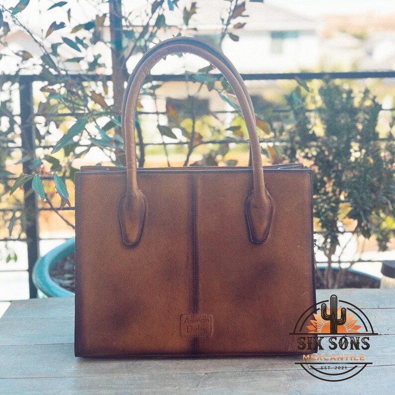 Puede incluir: Un bolso de mano estructurado de cuero marr&oacute;n con dos asas superiores y una costura vertical en el centro. El bolso presenta una silueta rectangular y una peque&ntilde;a etiqueta con el logotipo grabado cerca de la base que dice American Darling.