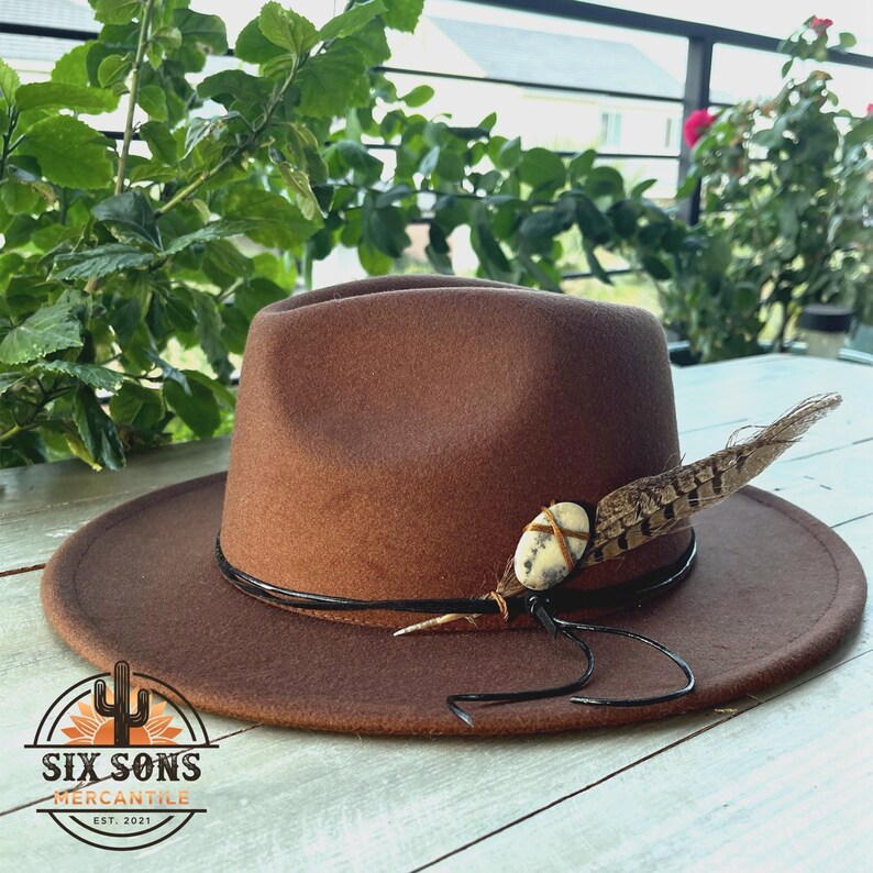 May include: A brown fedora hat with a black leather band and a feather accent. The hat has a stone and leather embellishment on the feather. The hat is labeled "Six Sons Mercantile Est. 2021".