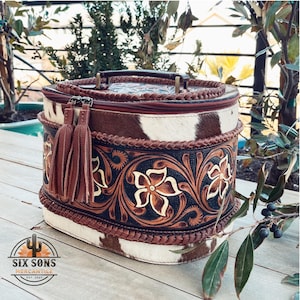 May include: A brown and white oval-shaped leather bag with a floral design and a braided handle. The bag has a brown tassel and a zipper closure. The bag is sitting on a wooden surface.