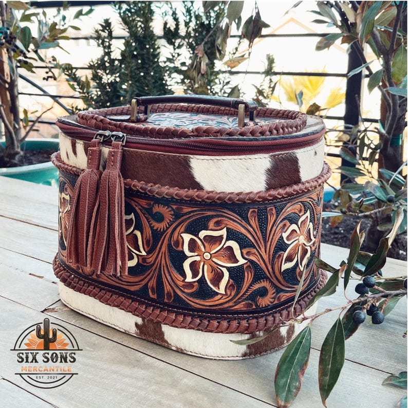 May include: A brown and white oval-shaped leather bag with a floral design and a braided handle. The bag has a brown tassel and a zipper closure. The bag is sitting on a wooden surface.