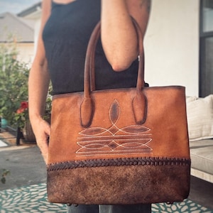 Puede incluir: Un bolso tote de cuero marr&oacute;n con dos asas superiores, un patr&oacute;n decorativo de costuras blancas en la secci&oacute;n superior y un borde de cuero trenzado marr&oacute;n oscuro que separa el cuerpo de una base de cuero texturizado m&aacute;s oscuro.