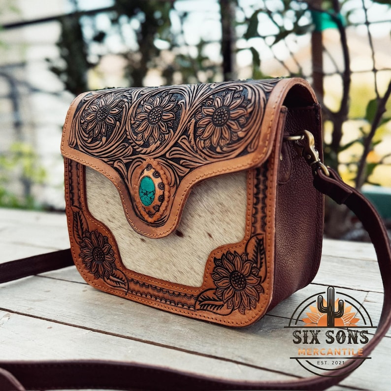 May include: A brown leather crossbody bag with a floral embossed design. The bag features a turquoise stone accent and a light-colored, textured front panel. The bag has a long brown strap and the words "SIX SONS MERCANTILE EST. 2021" are visible.
