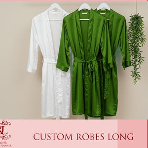 May include: Two long satin robes, one white and one green, hanging on hangers.  The robes have a shawl collar and tie belt.  "CUSTOM ROBES LONG" is written in red text at the bottom of the image.