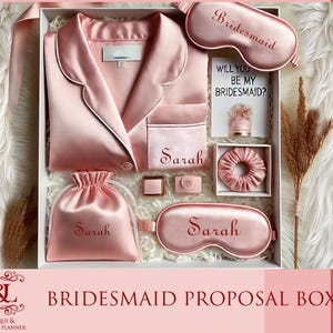 May include: A pink bridesmaid proposal box with a satin sleep mask, robe, and a card that says "Will you be my bridesmaid?" The box also includes a small satin bag, a hair scrunchie, and a ring box. The items are personalized with the name "Sarah".